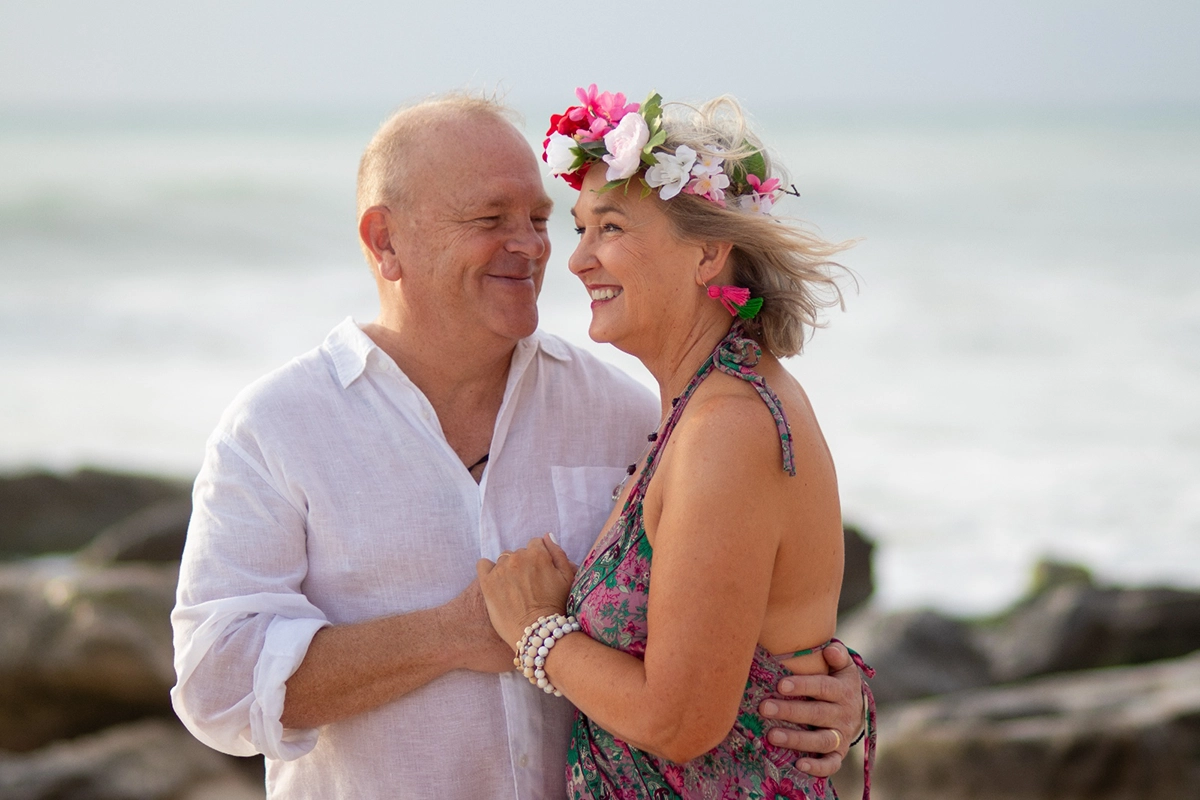 Pareja madura sonriendo y abrazándose en la playa; mujer con corona de flores rosas y vestido boho, hombre con camisa blanca de lino, con fondo de mar difuminado.