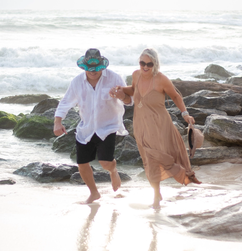 Pareja de adultos mayores sonriendo y corriendo de la mano en una playa tropical con olas y rocas de fondo.