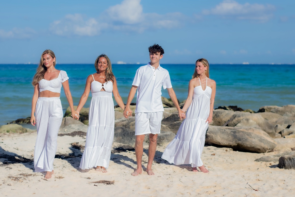 Familia de cuatro integrantes posando sonrientes y abrazados en la orilla de una playa tropical con palmeras y mar azul.