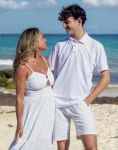 Retrato emotivo de una madre abrazando a su hijo pequeño en la orilla de una playa del Caribe, ambos vestidos de blanco con el mar de fondo.
