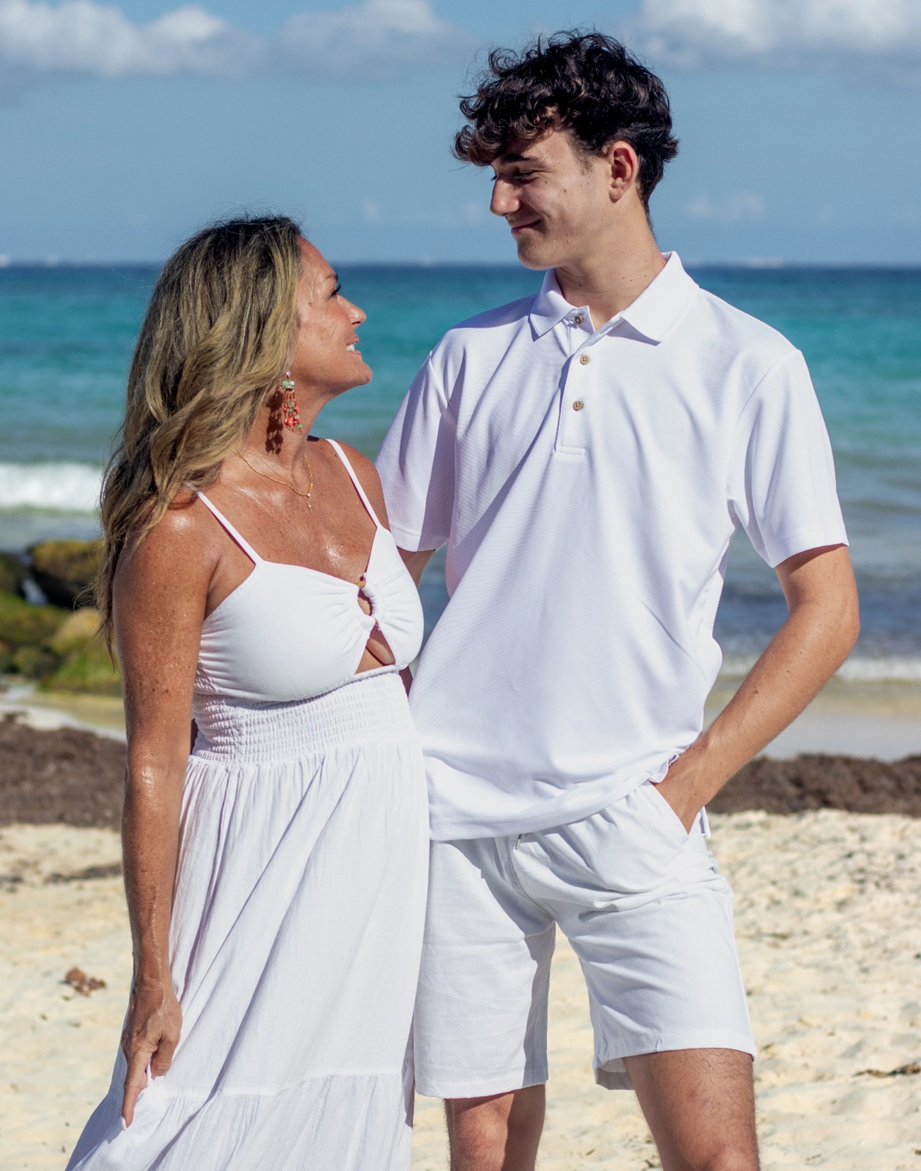 Retrato emotivo de una madre abrazando a su hijo pequeño en la orilla de una playa del Caribe, ambos vestidos de blanco con el mar de fondo.