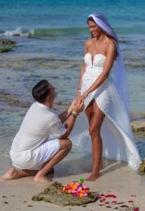 Hombre arrodillado pidiendo matrimonio a una mujer vestida de novia en una playa de aguas cristalinas.