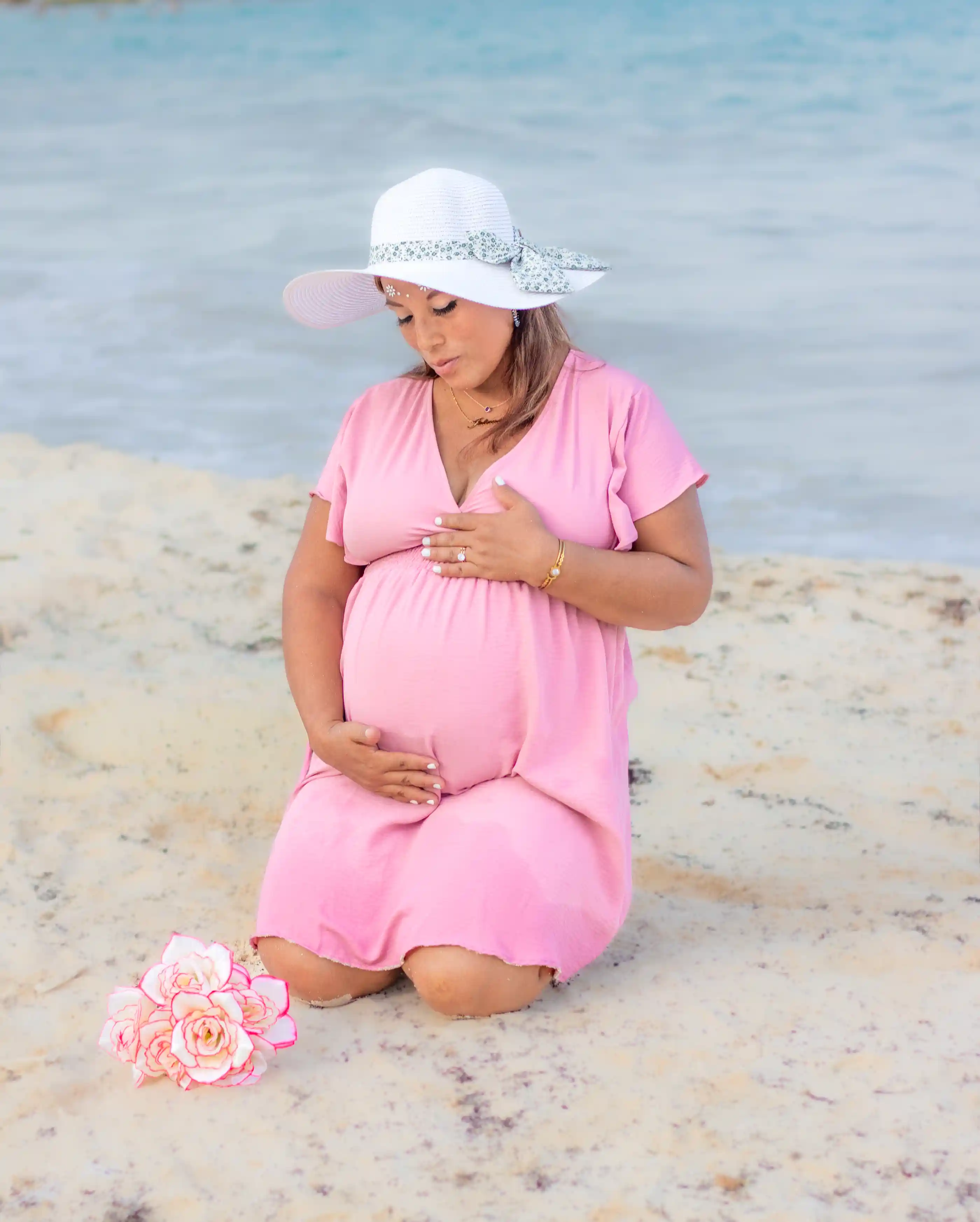 Pregnant woman kneeling on beach in pink dress – maternity photography in Playa del Carmen