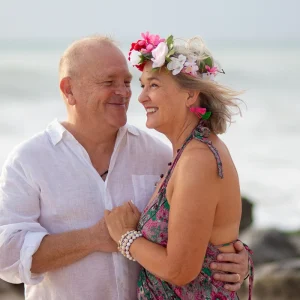 Pareja madura sonriendo y abrazándose en la playa; mujer con corona de flores rosas y vestido boho, hombre con camisa blanca de lino, con fondo de mar difuminado.